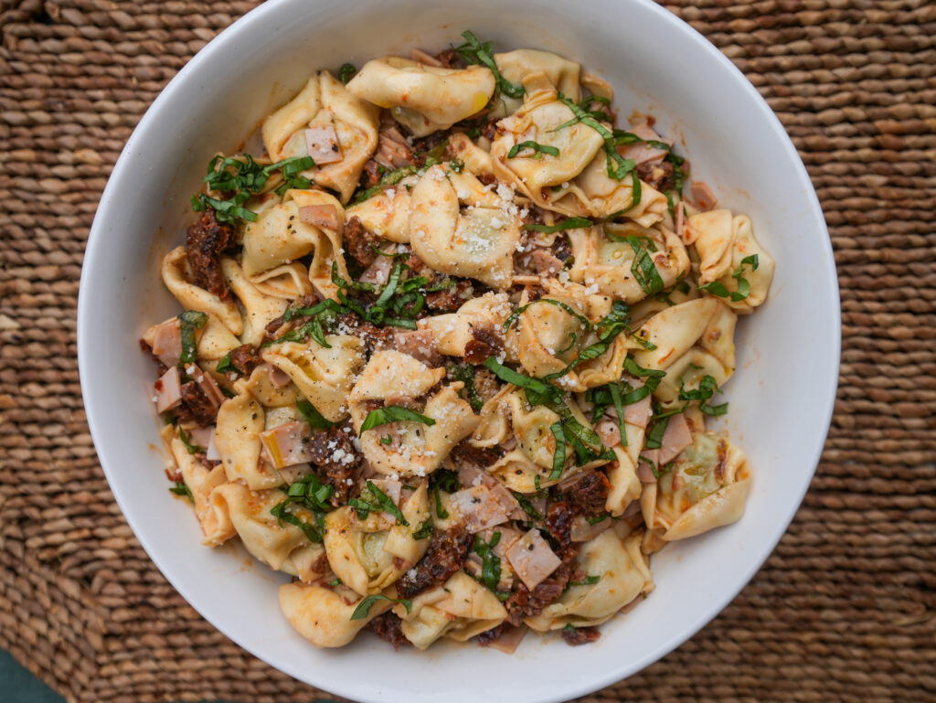 Bowl of Marry Me Tortellini Salad with mascarpone dressing, sun-dried tomatoes, mortadella, spinach, and fresh basil served cold