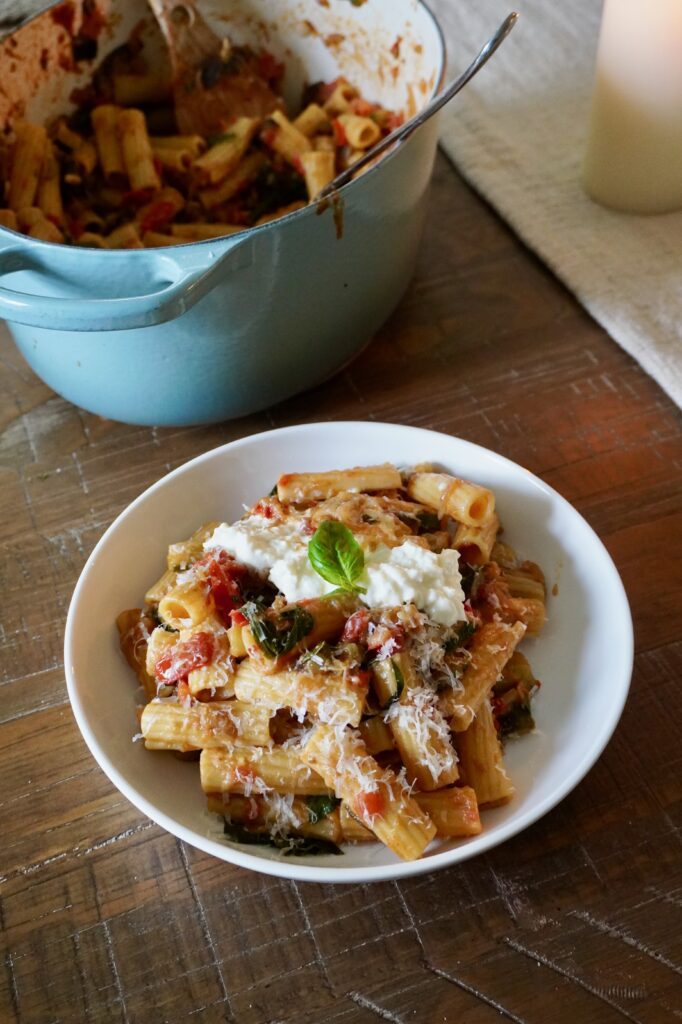Late summer vegetable pasta recipe featuring rigatoni with caramelized ratatouille sauce, topped with stracciatella, Parmesan, and basil on a rustic table