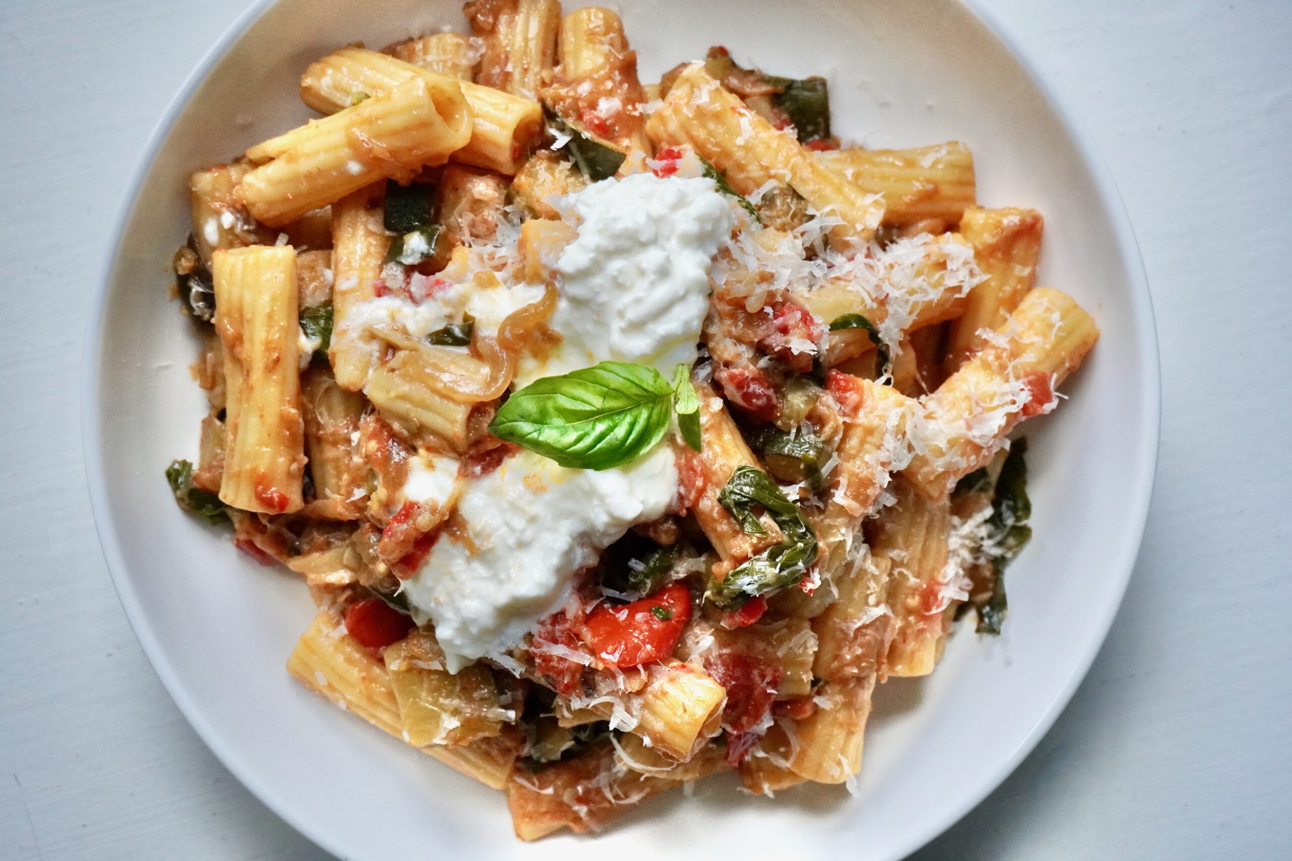 A bowl of rigatoni pasta coated in caramelized ratatouille sauce made with eggplant, zucchini, bell pepper, onion, and tomatoes, topped with stracciatella cheese and fresh basil