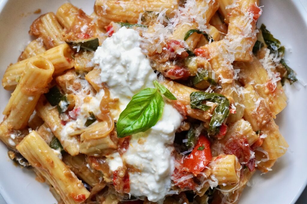 Close-up of rigatoni pasta with caramelized summer vegetables in a tomato-based ratatouille sauce, finished with creamy stracciatella and grated Parmesan