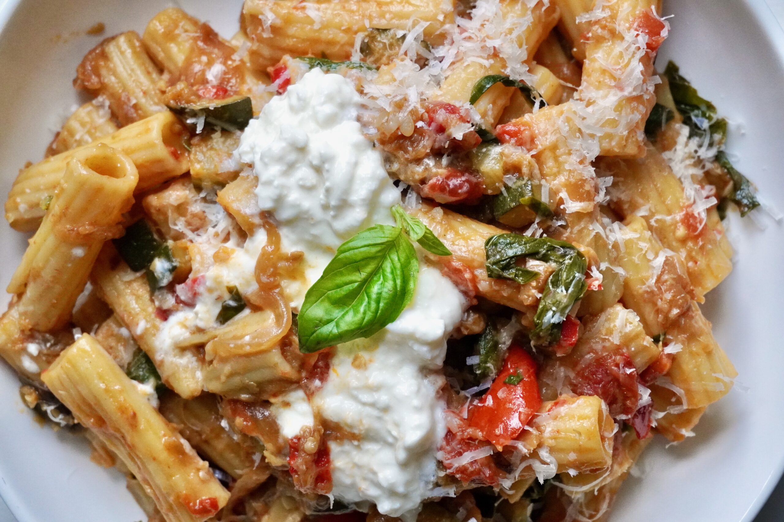 Close-up of rigatoni pasta with caramelized summer vegetables in a tomato-based ratatouille sauce, finished with creamy stracciatella and grated Parmesan