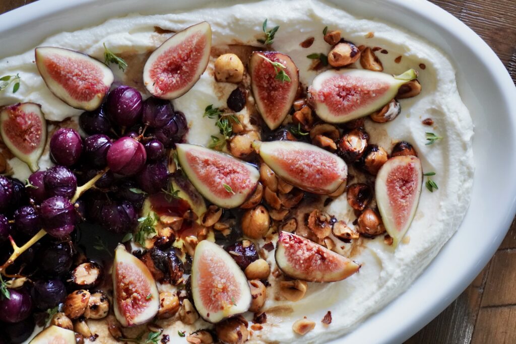 Overhead shot of whipped feta with roasted grapes, figs, and hazelnuts on a large oval platter, served with toasted baguette slices.