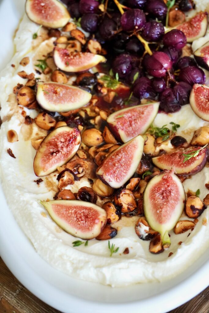 Close-up of glossy roasted grapes and fresh fig quarters arranged over a creamy whipped feta base, sprinkled with toasted hazelnuts and thyme.