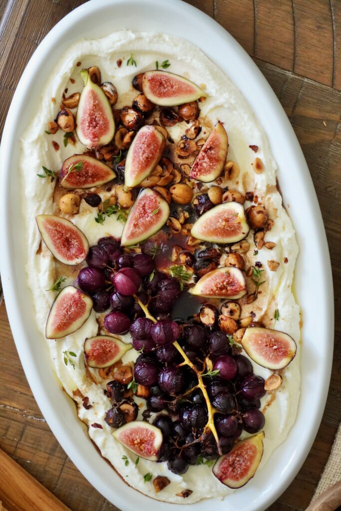 Overhead shot of whipped feta with roasted grapes, figs, and hazelnuts on a large oval platter, served with toasted baguette slices. shot in vertical angle.