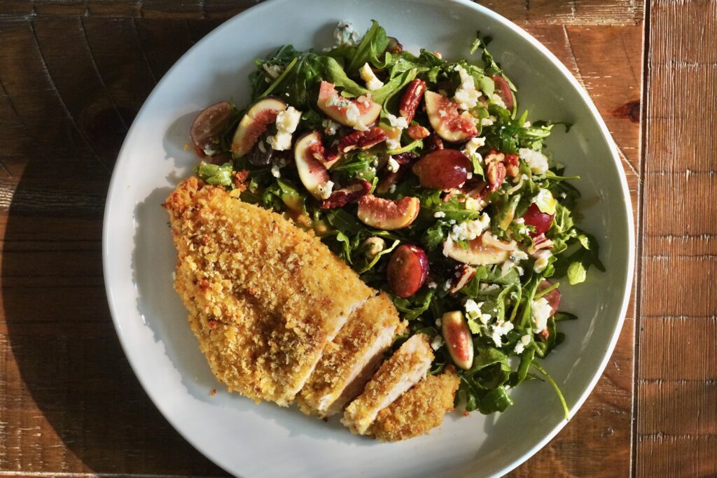 Crispy baked chicken cutlets sliced and served with arugula, red grapes, toasted pecans, shallots, and crumbled blue cheese on a rustic plate.