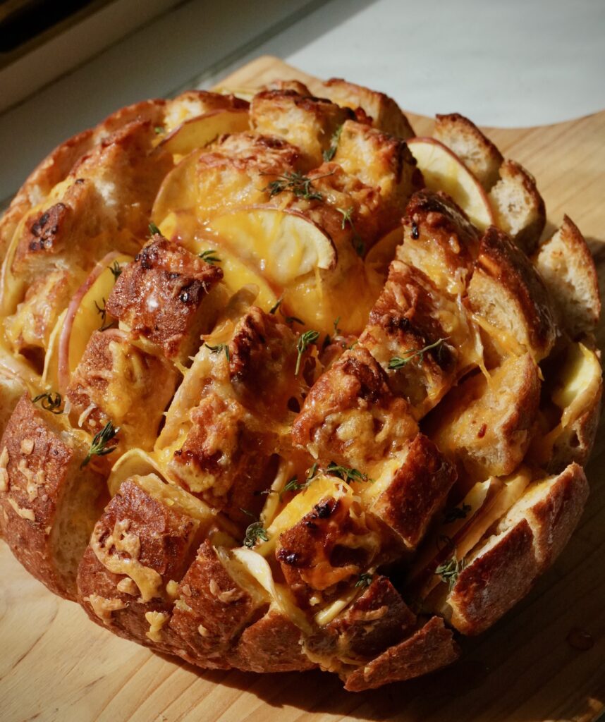 Golden sourdough pull apart bread stuffed with melted cheddar cheese and apple slices, drizzled with hot honey and topped with fresh thyme in the sunshine