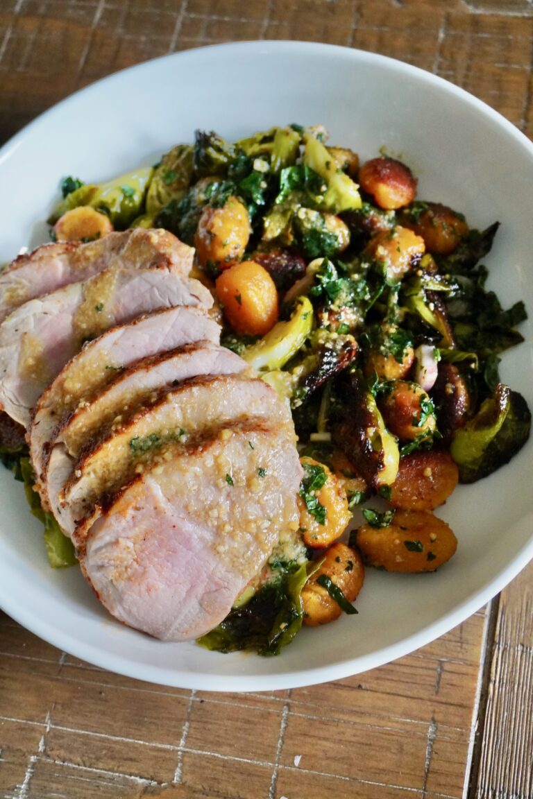 Close-up of a bowl filled with sliced pork tenderloin, roasted pumpkin gnocchi, caramelized Brussels sprouts, and wilted kale drizzled with white miso vinaigrette.