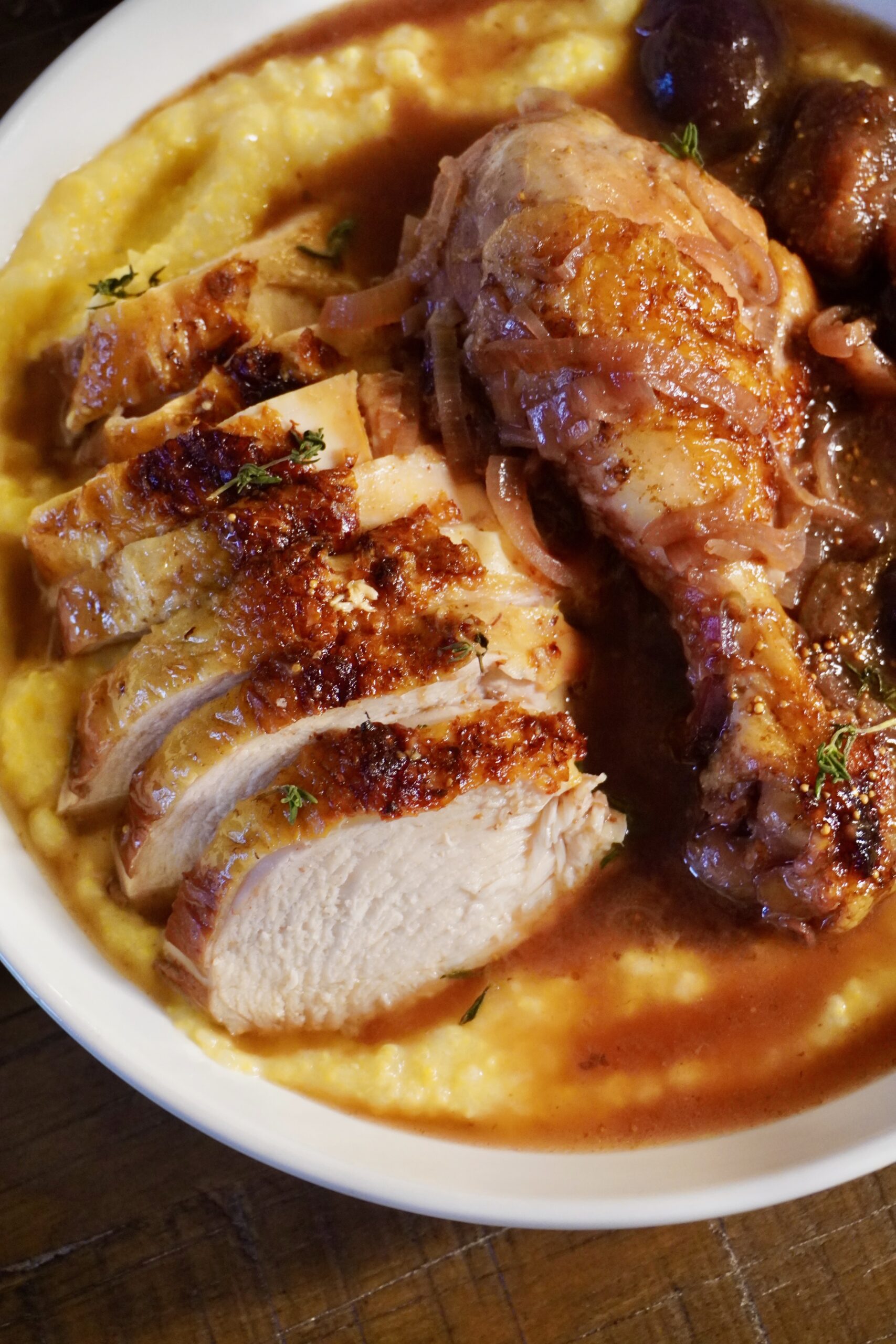 A rustic ceramic bowl filled with braised chicken pieces and figs in a glossy red wine balsamic sauce served over creamy polenta.