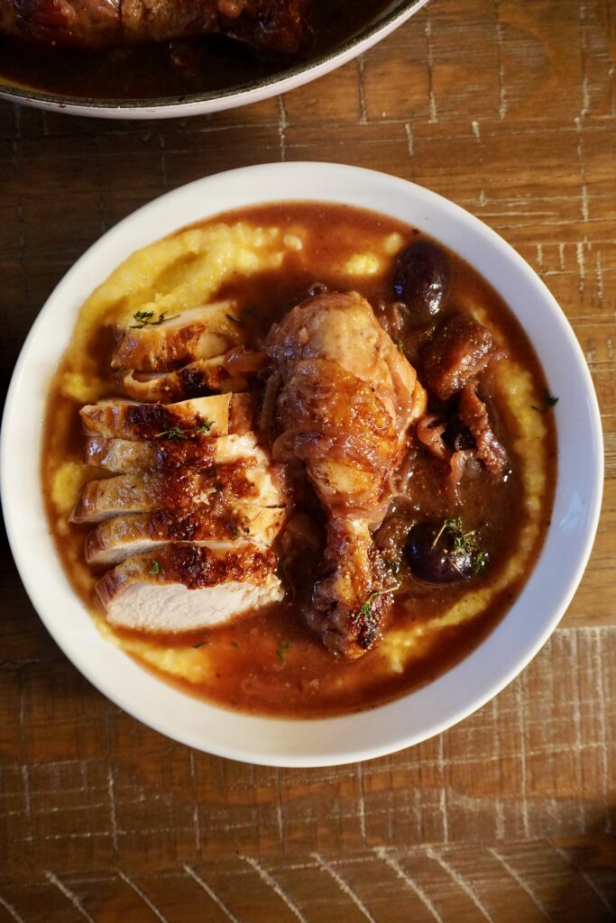 Overhead shot of a single plate with braised chicken, figs, and red wine sauce spooned over polenta, garnished with fresh thyme.