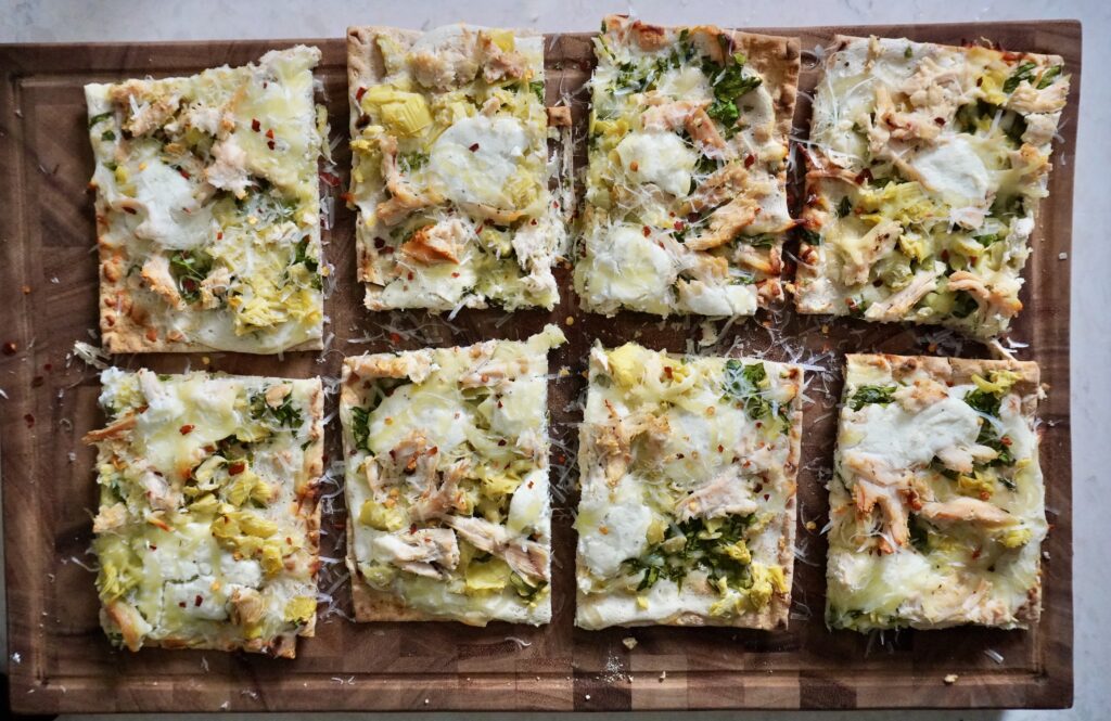 Close up of a crispy flatbread slice showing bubbling golden cheese, tender chicken chunks, and creamy spinach artichoke topping
