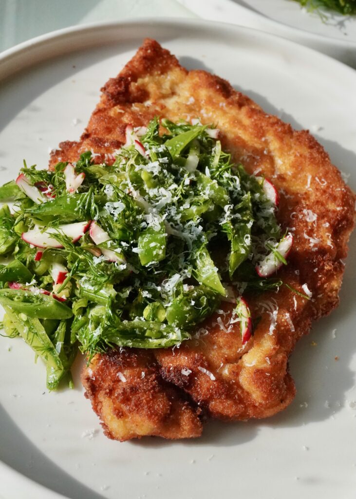 A close-up of the shatteringly crisp edge of a fried chicken cutlet topped with a vibrant green snap pea and radish salad