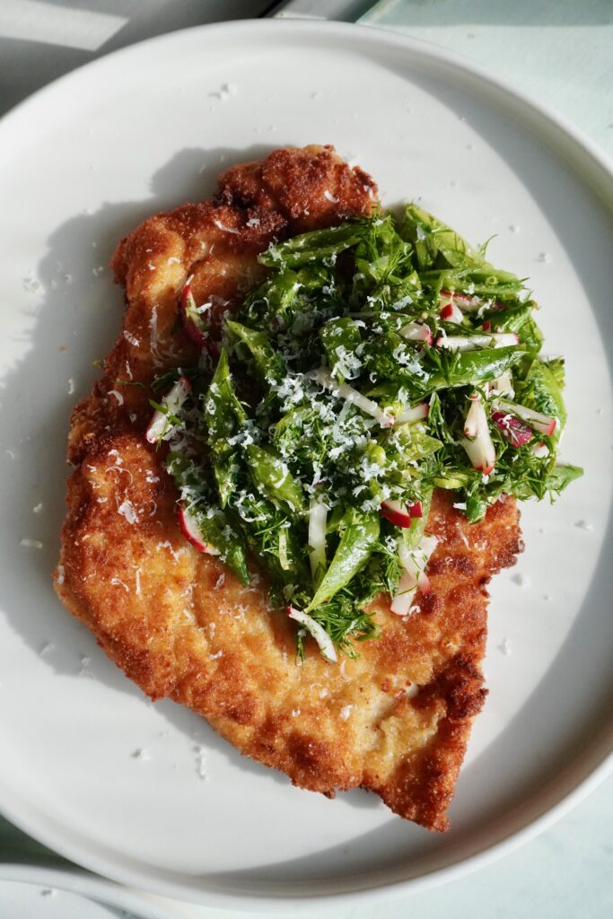A golden, crispy chicken cutlet topped with a vibrant green snap pea and radish salad