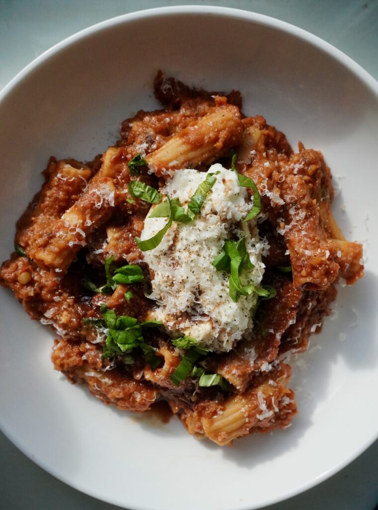 A bowl of rigatoni pasta coated in a thick eggplant and lentil ragu, topped with a ball of torn burrata