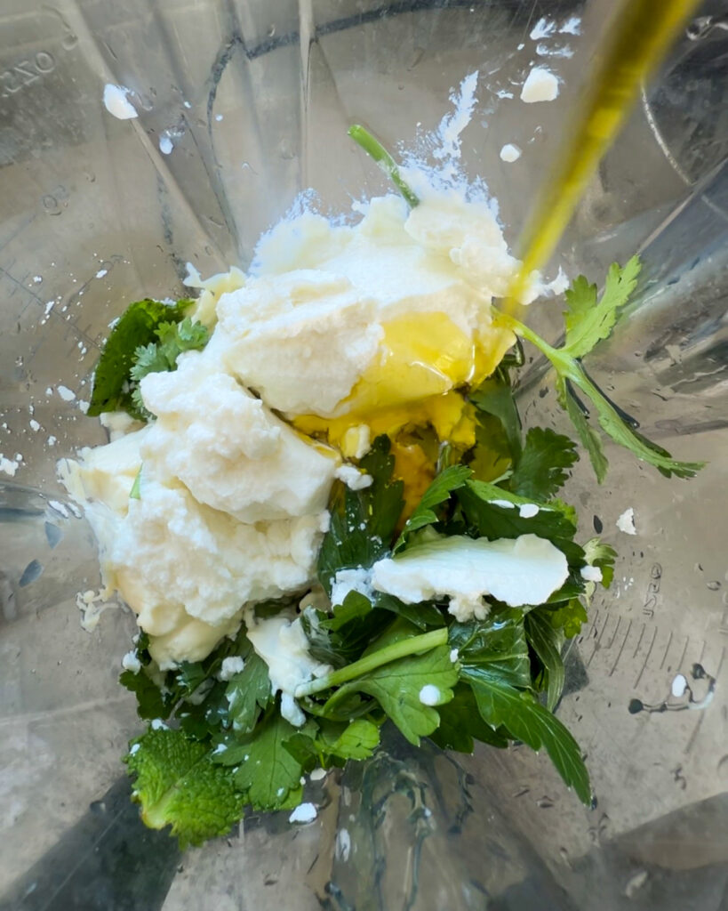 Fresh parsley, cilantro, and mint being added to a blender with Greek yogurt and miso