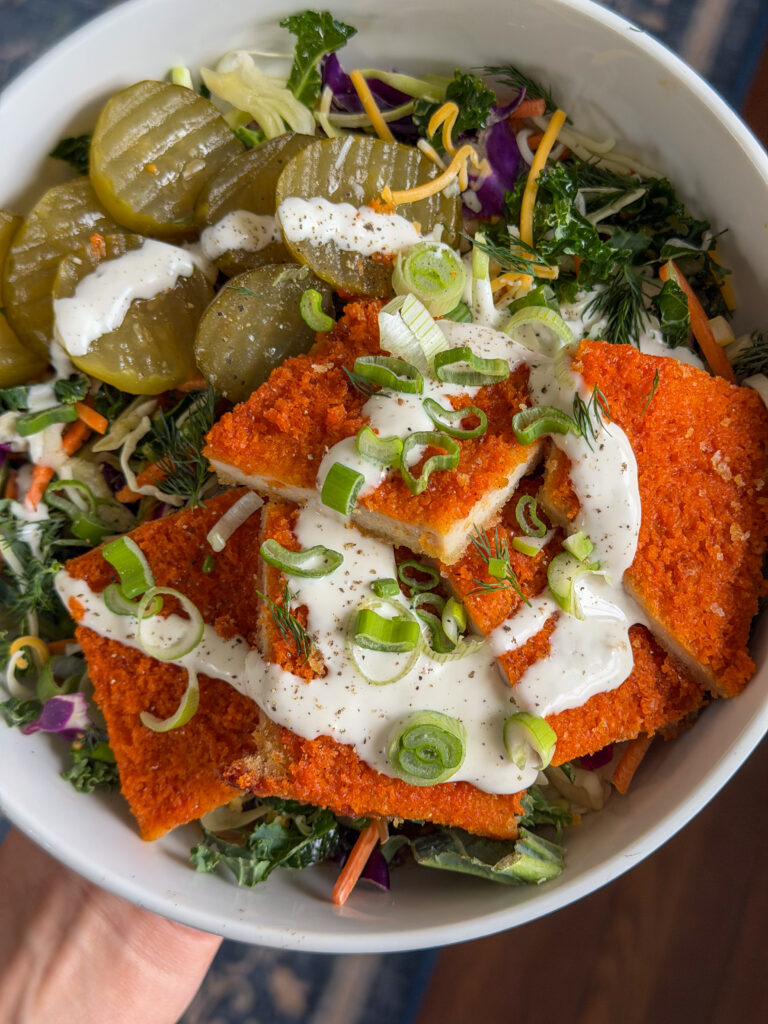 Close-up of golden oven-fried crispy Nashville hot chicken cutlet sliced over fresh slaw with creamy ranch drizzle, pickles and herbs