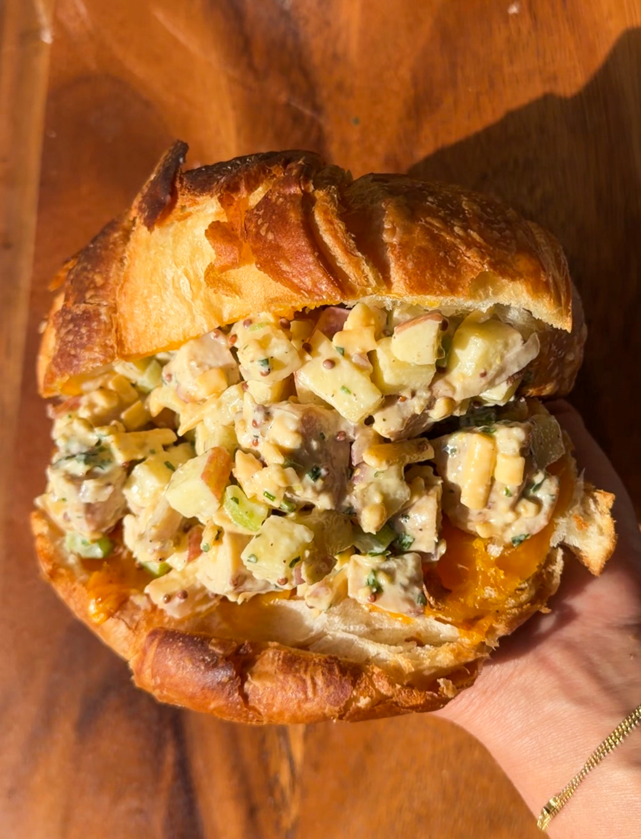 Apple cheddar chicken salad piled into a toasted croissant with melted sharp cheddar, held over a wooden board
