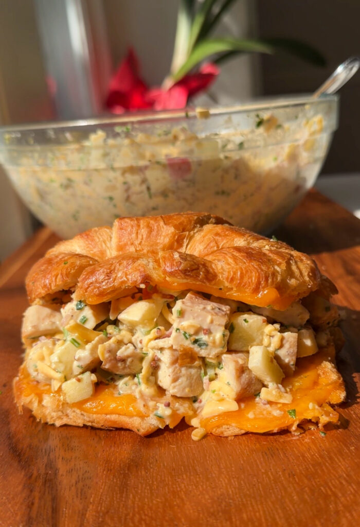 Apple cheddar chicken salad croissant melt with melted cheddar on a wooden board, with a bowl of chicken salad behind it