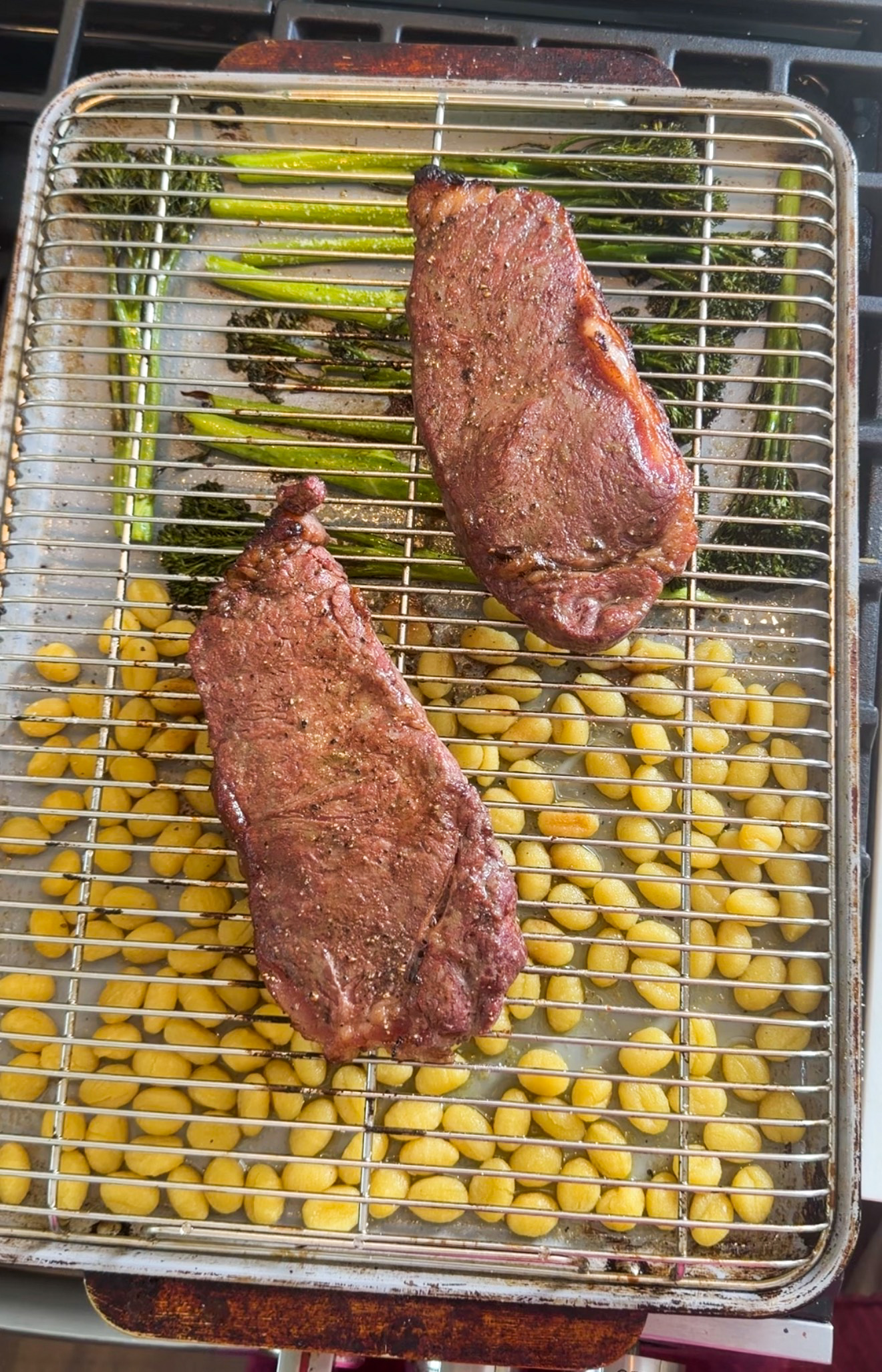Two whole strip steaks on a wire rack above roasted mini gnocchi and broccolini on a sheet pan