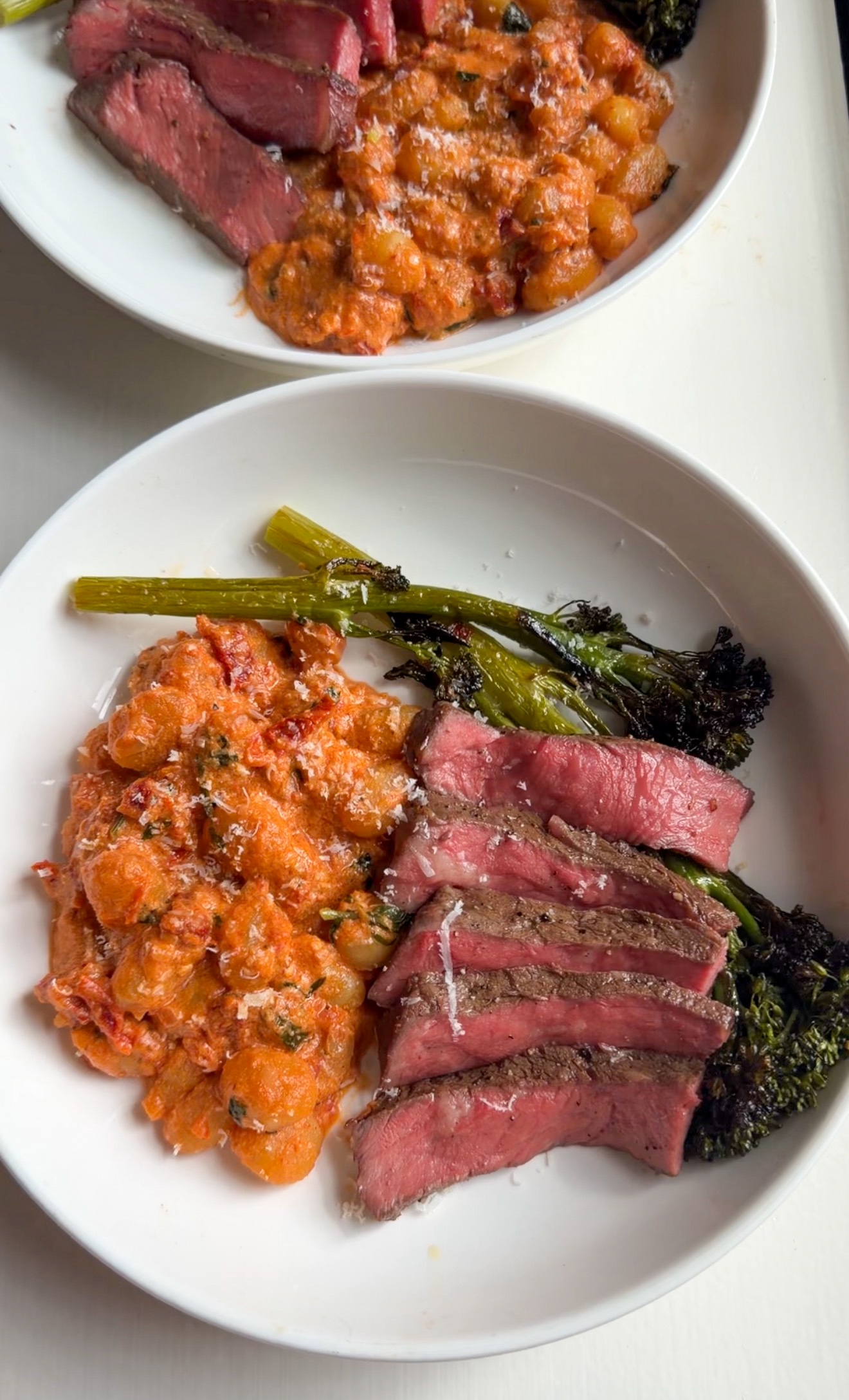 Two plates of sliced strip steak, creamy tomato gnocchi, and roasted broccolini with grated Parmesan