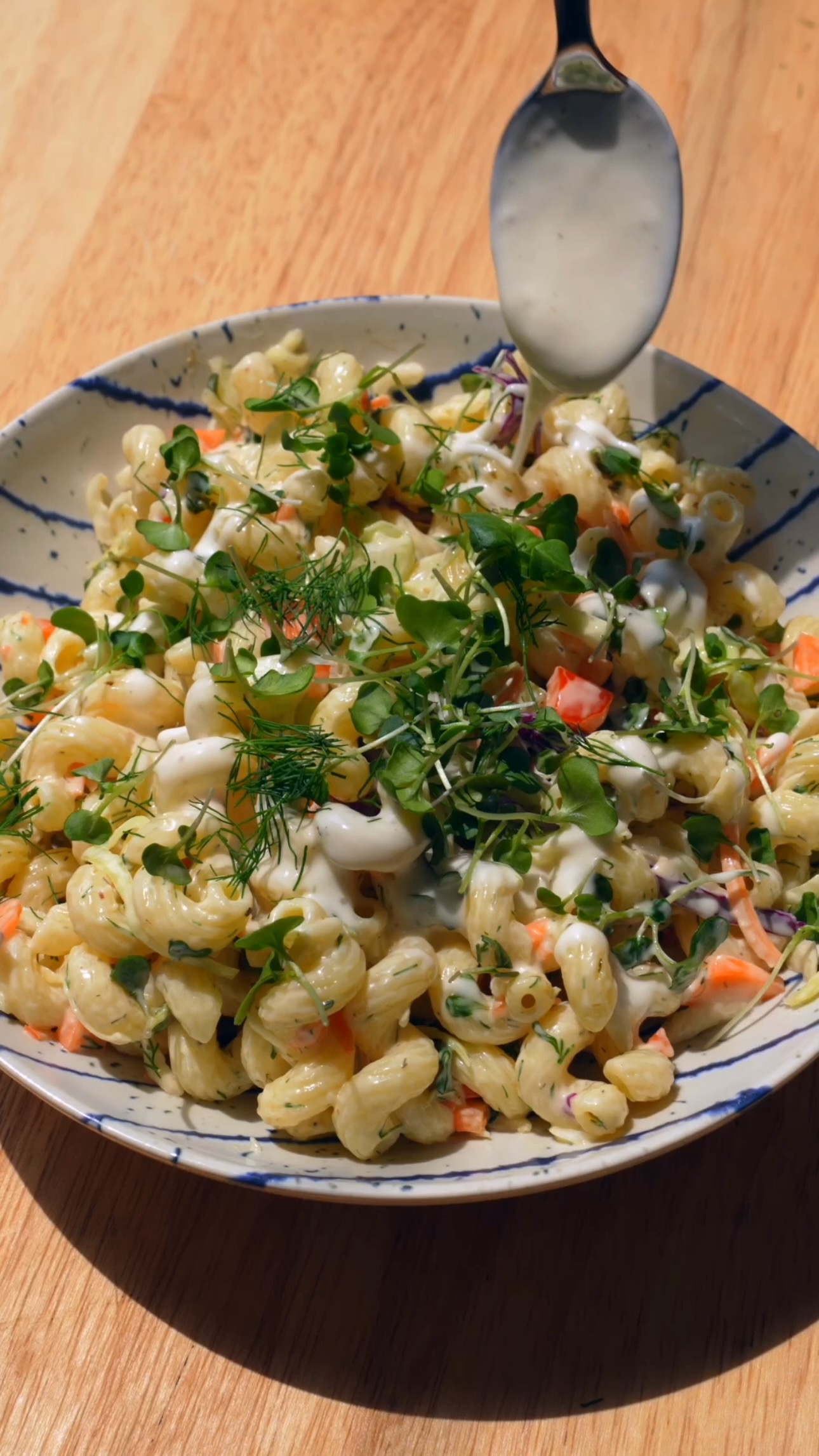 Ranch dressing being drizzled over cavatappi pasta salad with vegetables and microgreens