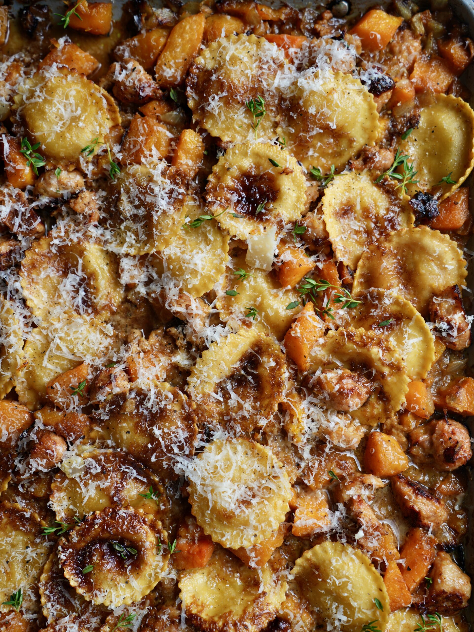Golden ravioli with roasted butternut squash and sausage on a sheet pan, scattered with Parmesan and fresh thyme.