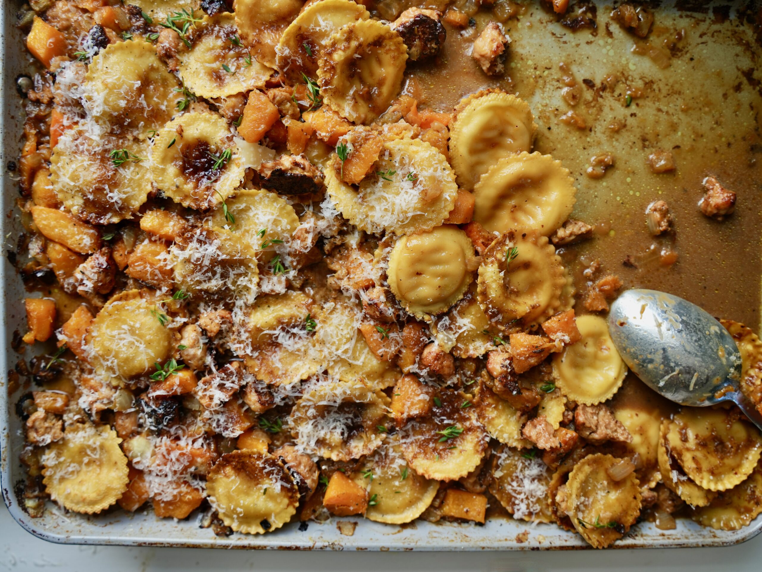 Sheet pan ravioli with sausage, roasted butternut squash, Parmesan and thyme, with a serving spoon resting on the pan.