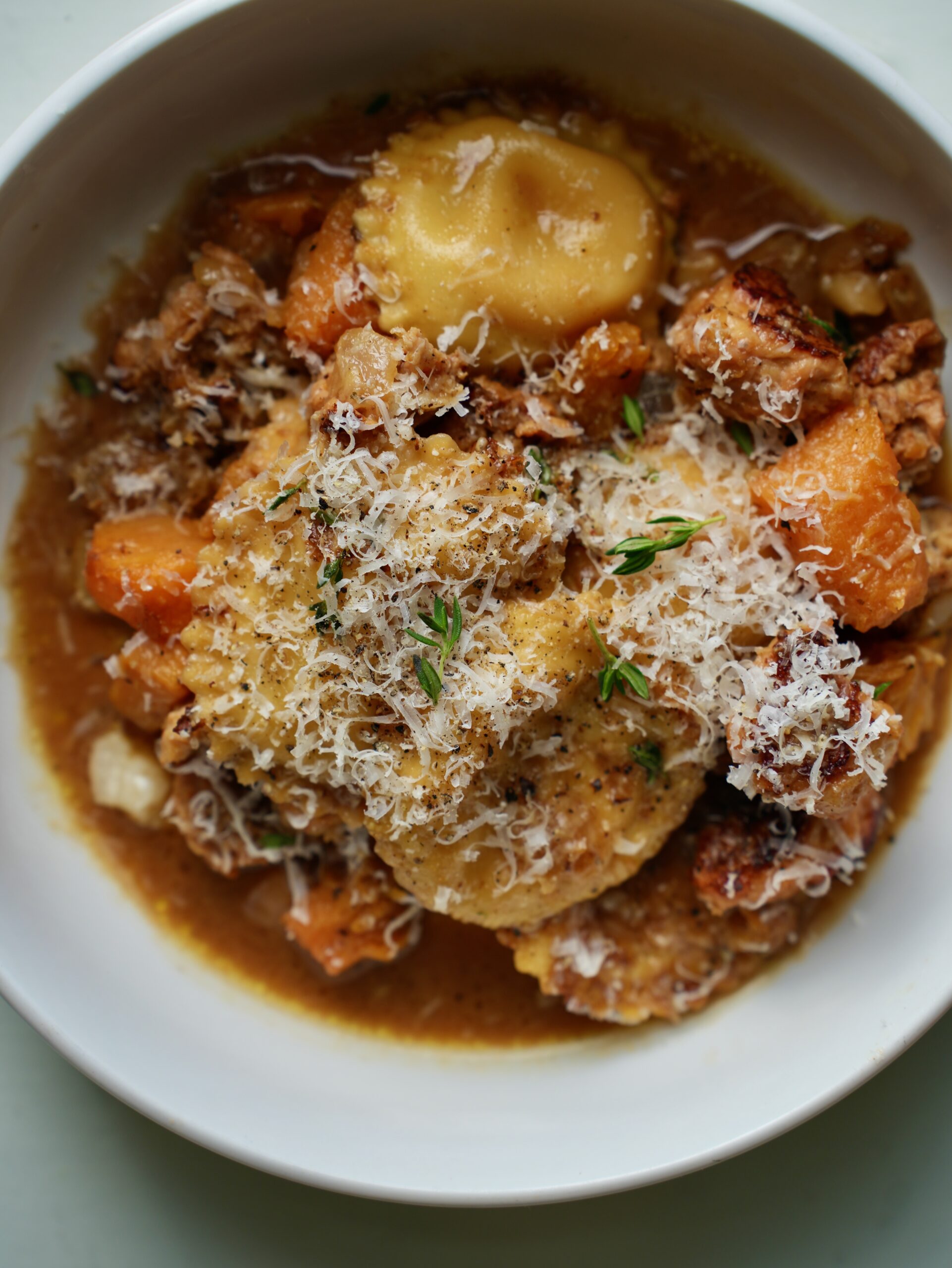 Ravioli, sausage and roasted butternut squash in creamy sauce served in a white bowl, topped with Parmesan and thyme.